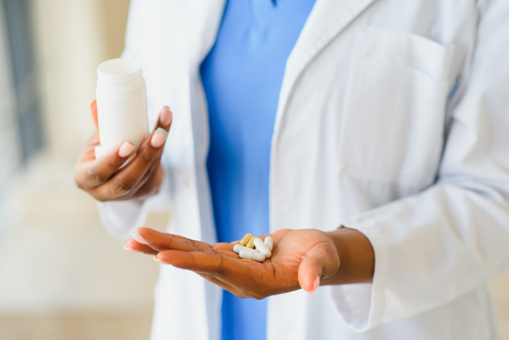 A person in a white lab coat holds a bottle in one hand and several pills and capsules in the other, illustrating a Sample Post about pharmaceutical care.