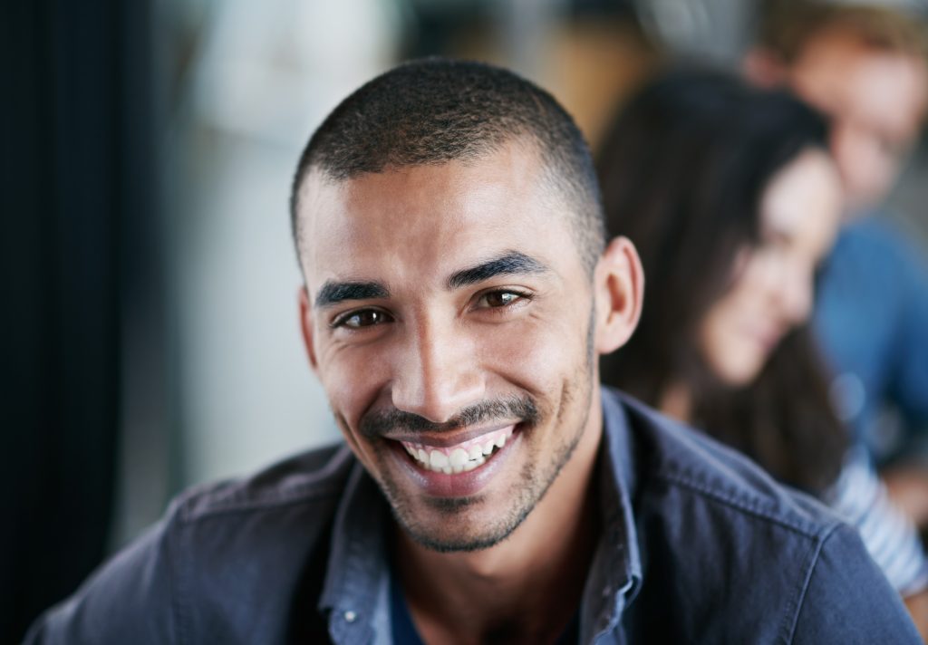 A man with short hair and facial hair smiles at the camera, with two people sitting blurred in the background in this Sample Post.