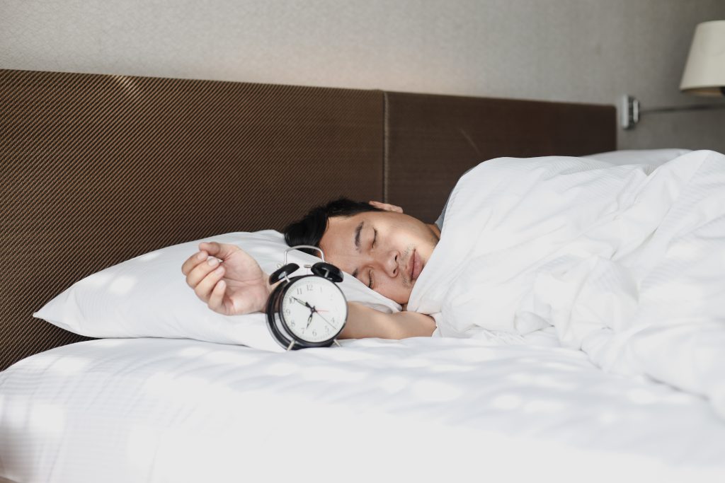 A person is sleeping in bed with their head on a pillow, covered by a white blanket, and an alarm clock resting near their hand on the pillow in this Sample Post.