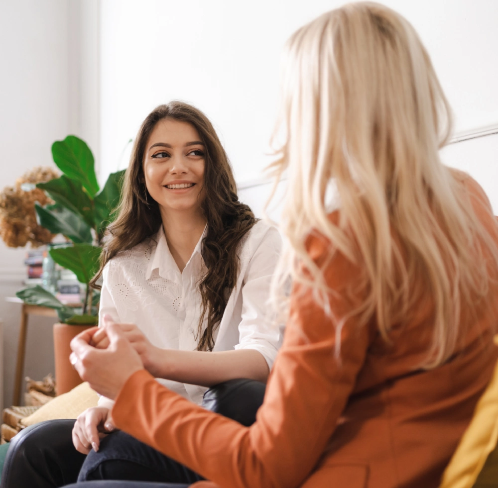 Two women sitting on a couch, engaged in conversation. One with long dark hair and white shirt, the other with blonde hair and an orange jacket. Indoor setting with plant in the background.