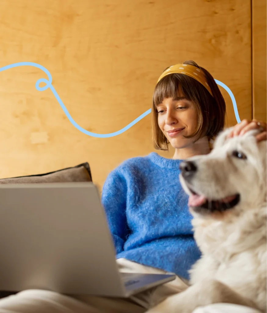 Person in blue sweater using a laptop, sitting on a sofa with a large white dog. Brown wooden wall in the background.