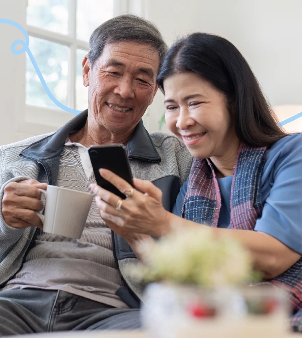 An older couple sits on a couch, smiling while looking at a smartphone; one person holds a coffee mug.