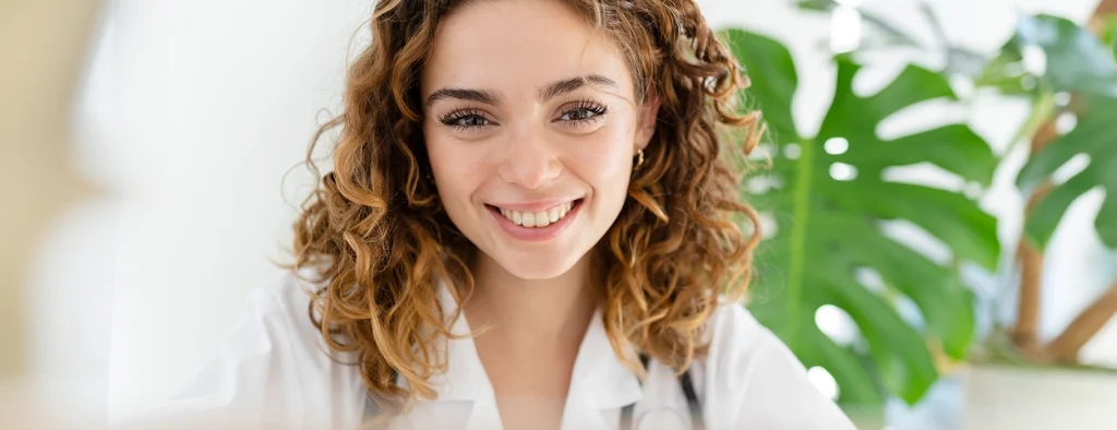 A young woman with curly hair, wearing a white coat, smiles at the camera with a green leafy plant in the background.
