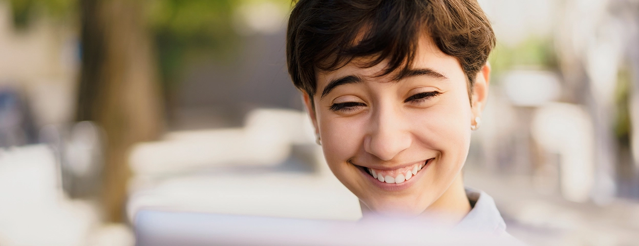 A person with short brown hair smiles while looking at a computer screen outdoors. Trees and sunlight are visible in the blurred background.