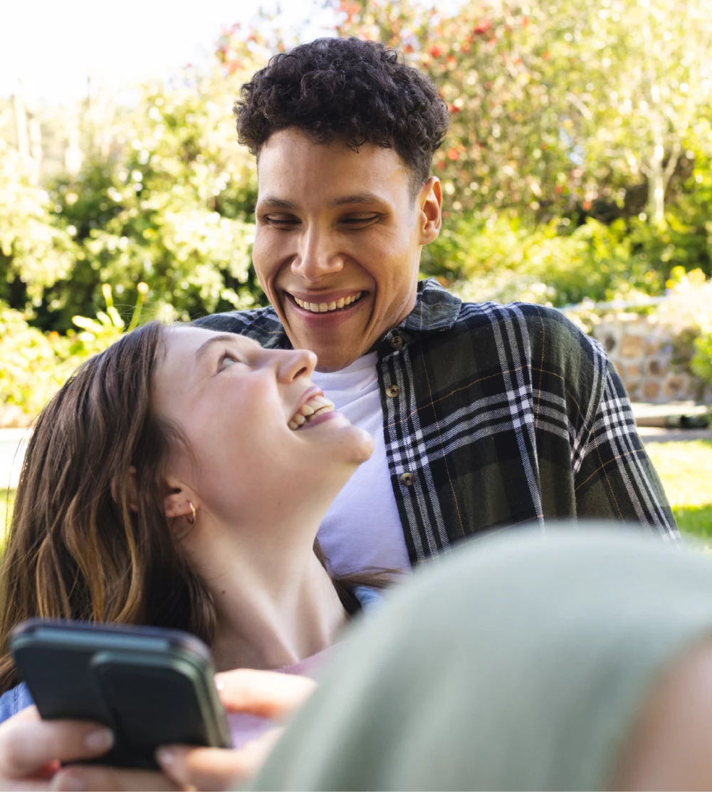 Two people sitting outdoors, smiling at each other, with one holding a smartphone. Lush greenery and flowers are visible in the background.