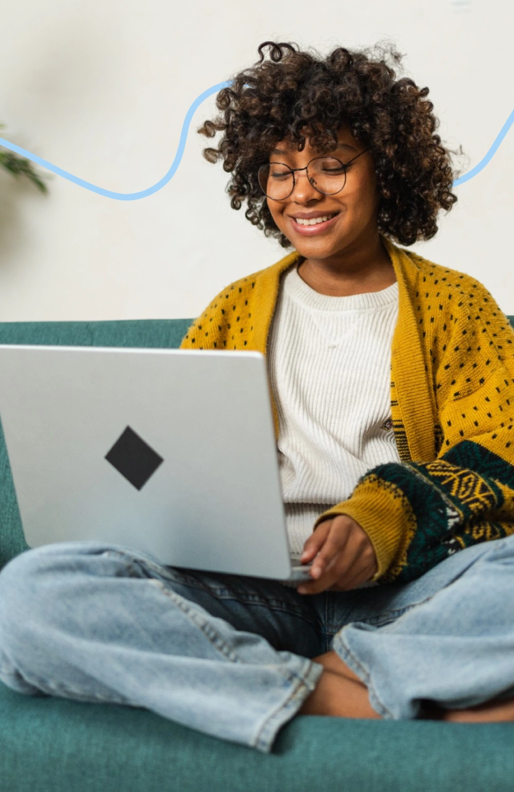 Person with curly hair and glasses, wearing a yellow cardigan, sitting cross-legged on a teal sofa, using a laptop.