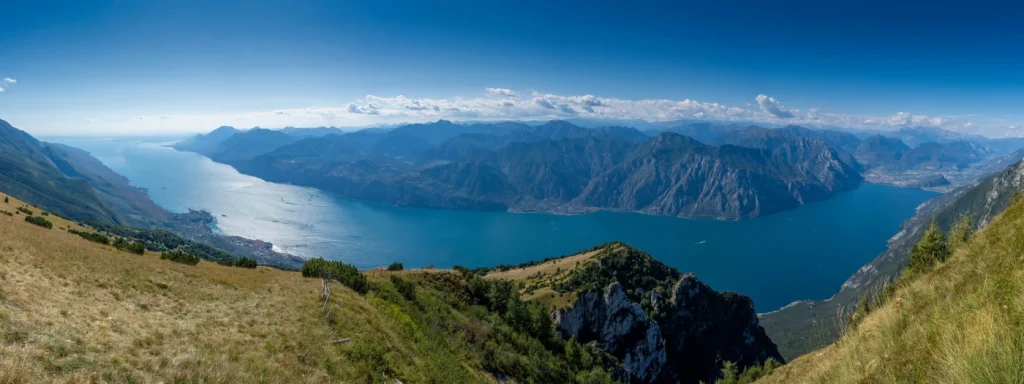 Panoramic view of a lake surrounded by mountains under a clear blue sky, with grassy hills in the foreground and distant towns along the shoreline.